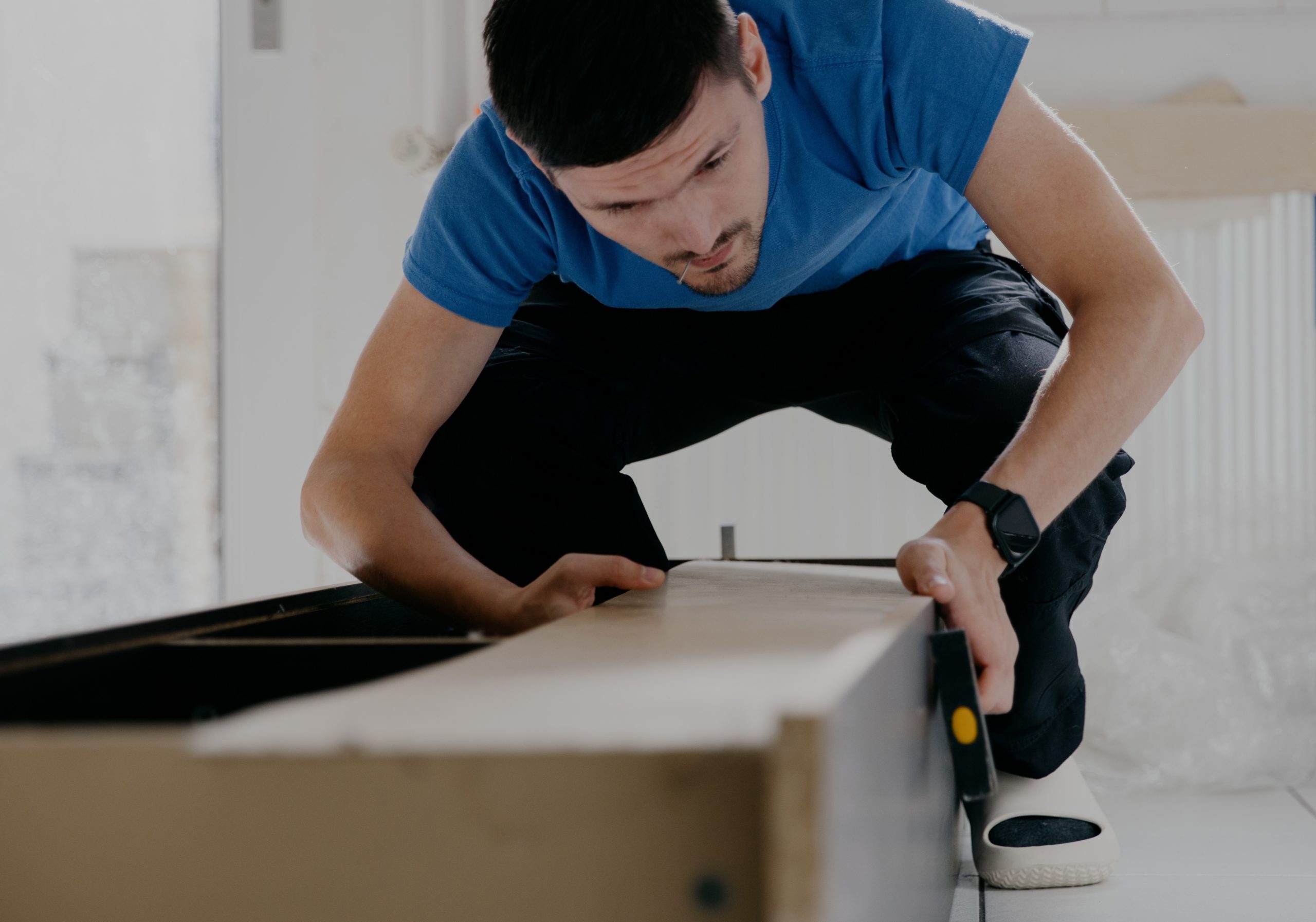 A man in a blue shirt assembles furniture, concentrating while holding a piece of the wooden frame and using a screwdriver.