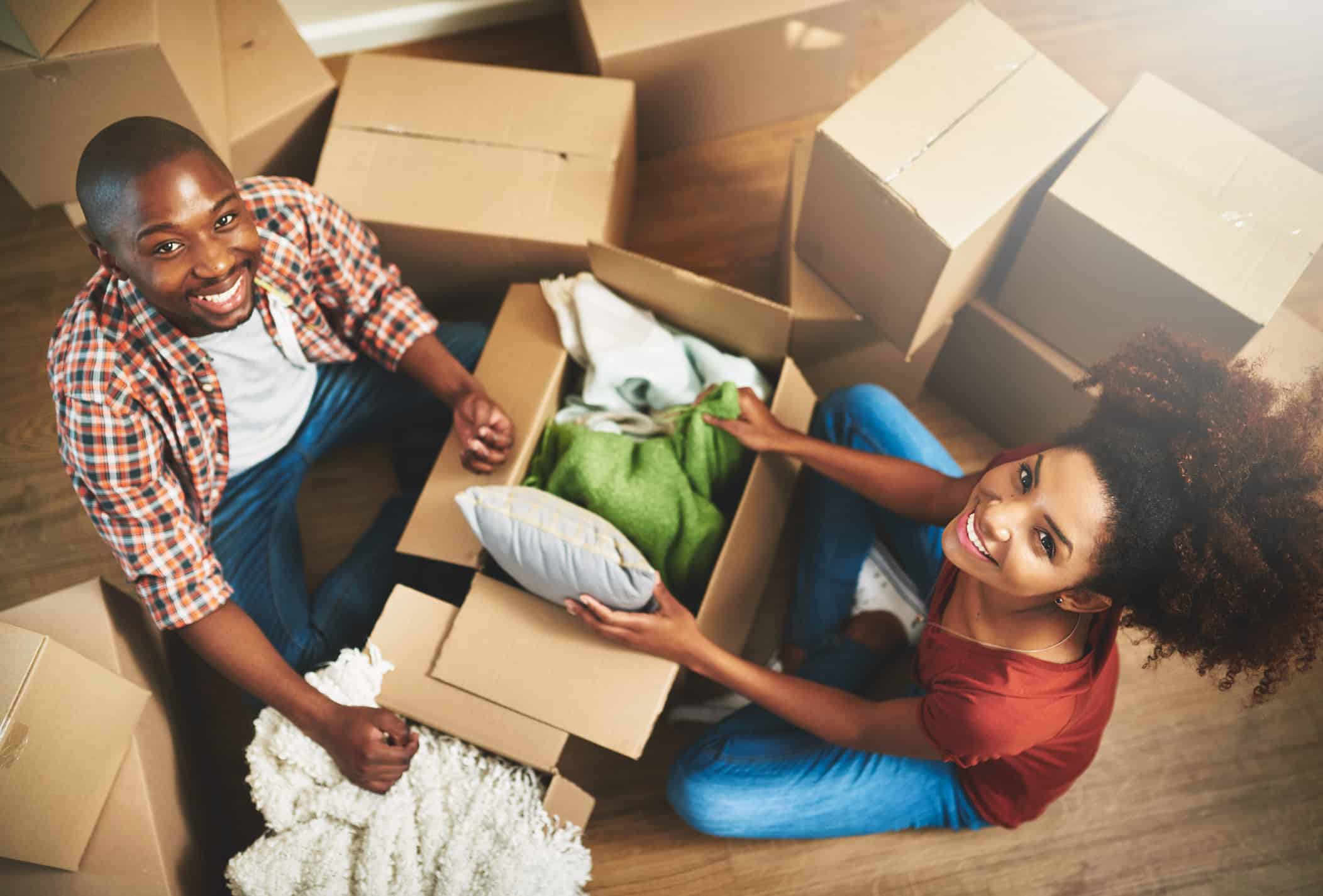 A cheerful couple unpacking boxes in their new home, sitting on the floor surrounded by cardboard boxes, looking up and smiling.